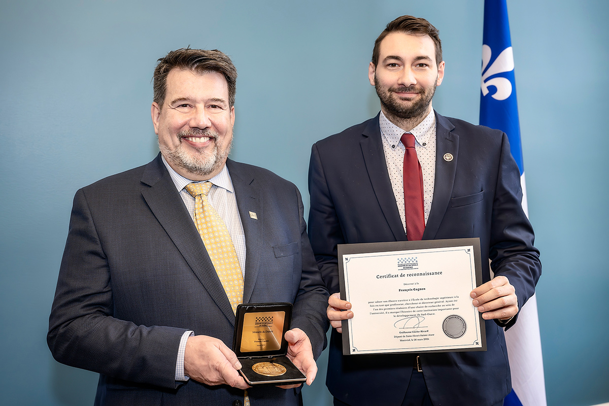 Remise de prix à un homme avec un certificat et médaille, souriants, drapeau québécois en arrière-plan.