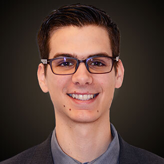 A confident young professional with glasses, wearing a suit, showcasing a warm, approachable smile.