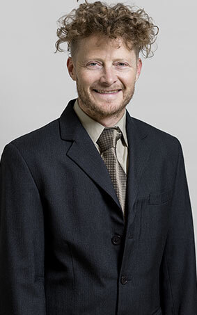 Homme souriant en costume sombre avec cravate à motif, cheveux bouclés. Représente un aspect professionnel.