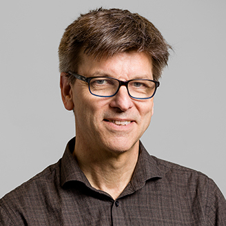 Homme souriant avec lunettes, chemise marron, fond gris, ambiance professionnelle et académique.