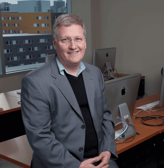 Professeur en costume devant des ordinateurs, souriant dans un bureau éclairé.