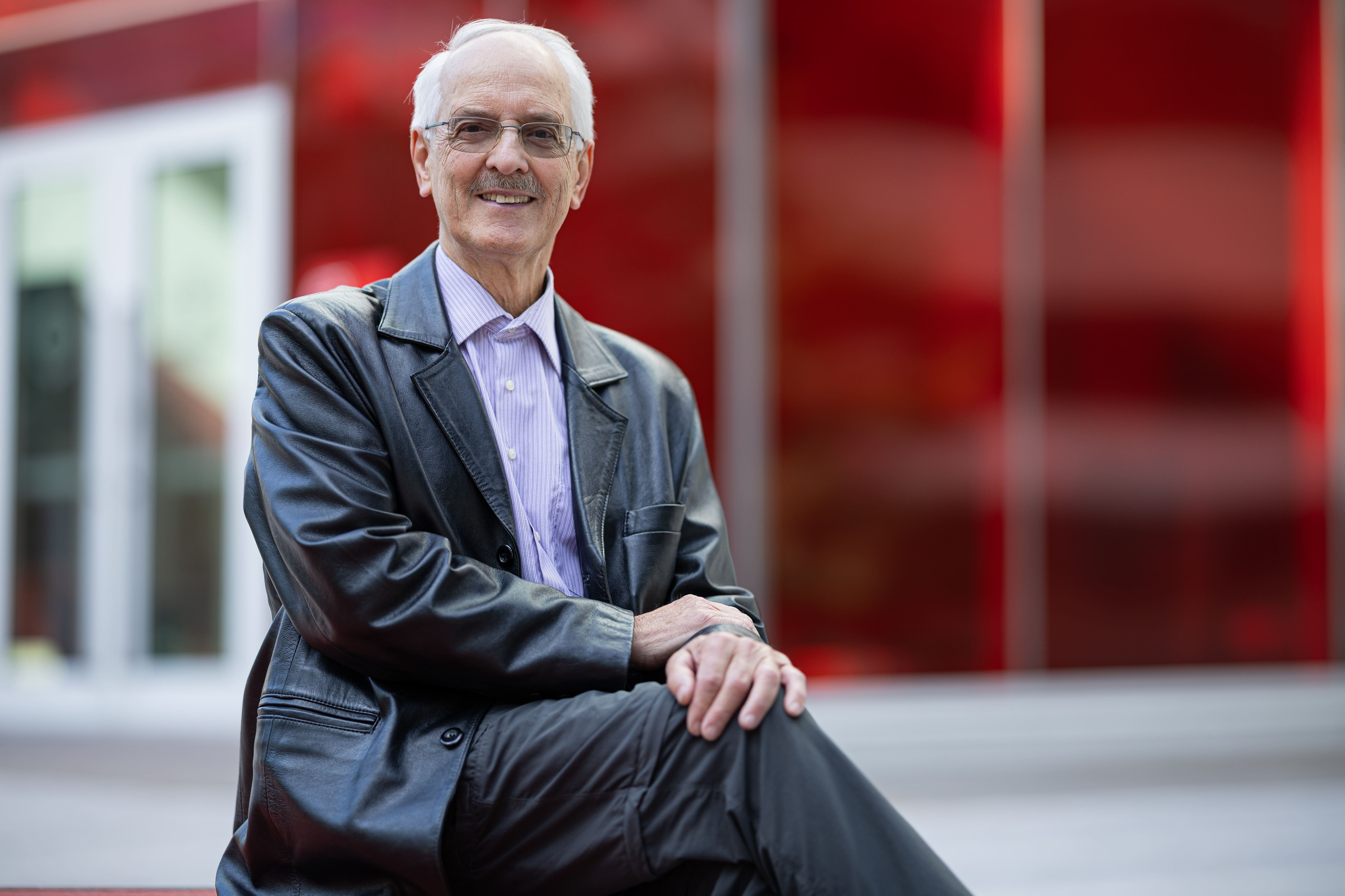 Professeur vétéran en technologie, souriant, assis devant un fond rouge moderne.