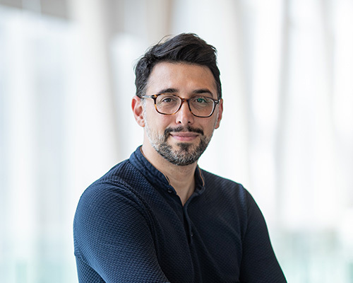 Confident academic with glasses and a beard in a casual sweater, posing in a modern university setting.