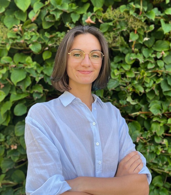 Étudiante en technologie souriante avec chemise bleue et lunettes, sur fond de verdure.