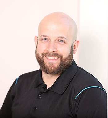 Un enseignant souriant, chauve, avec barbe, vêtu d'une chemise noire.