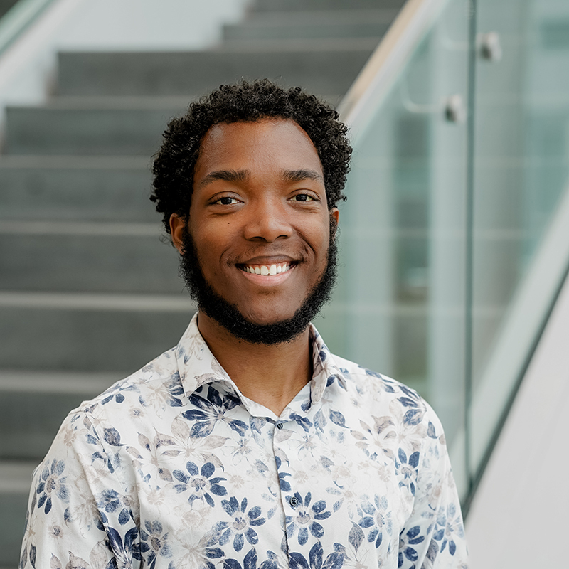 Étudiant souriant, chemise à fleurs, devant escaliers en verre, incarne la diversité en technologie.