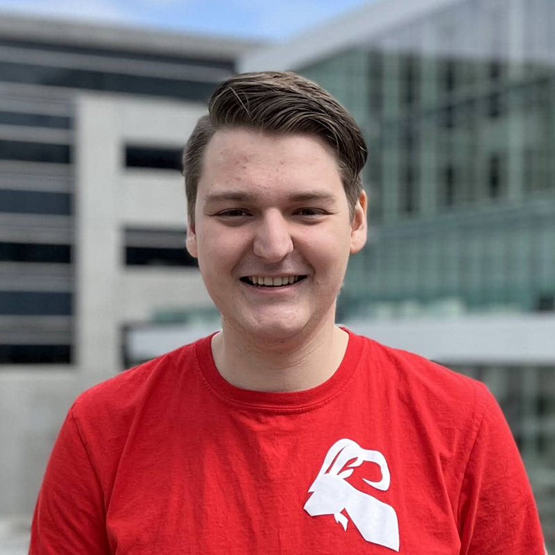 Étudiant souriant en tee-shirt rouge devant un bâtiment universitaire.