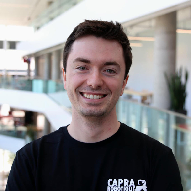 Étudiant souriant, portant un t-shirt de Capra Robotique, dans un hall universitaire.