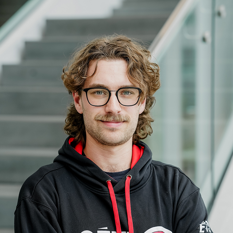 Étudiant souriant avec lunettes, sweat à capuche noir, devant un escalier.