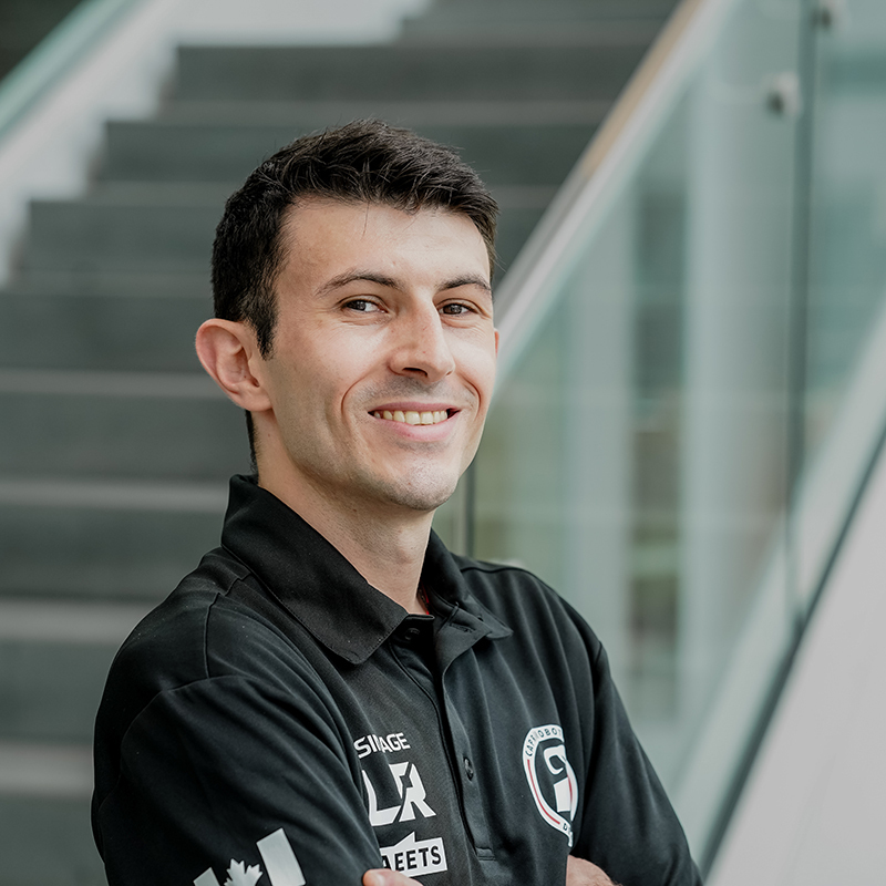 Étudiant souriant en technologie, portant un polo avec logos, devant un escalier.