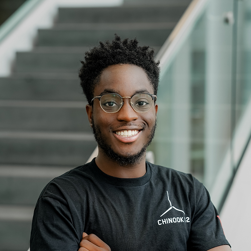 Étudiant souriant, vêtu d'un t-shirt noir, avec des lunettes, dans un milieu universitaire.