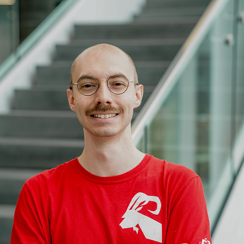 Étudiant souriant en t-shirt rouge, posant devant un escalier en verre.