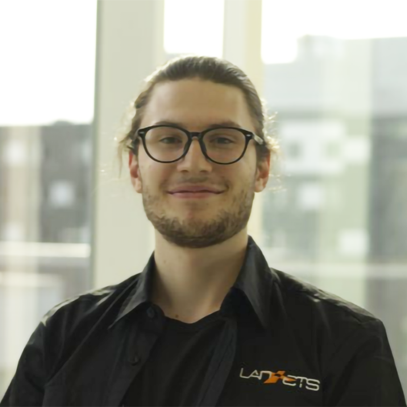 Étudiant souriant avec lunettes, chemise noire et logo universitaire.