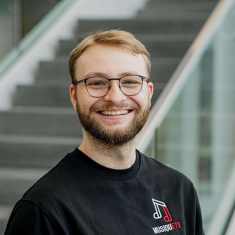 Étudiant souriant avec des lunettes, vêtu d'un T-shirt noir dans un bâtiment moderne.