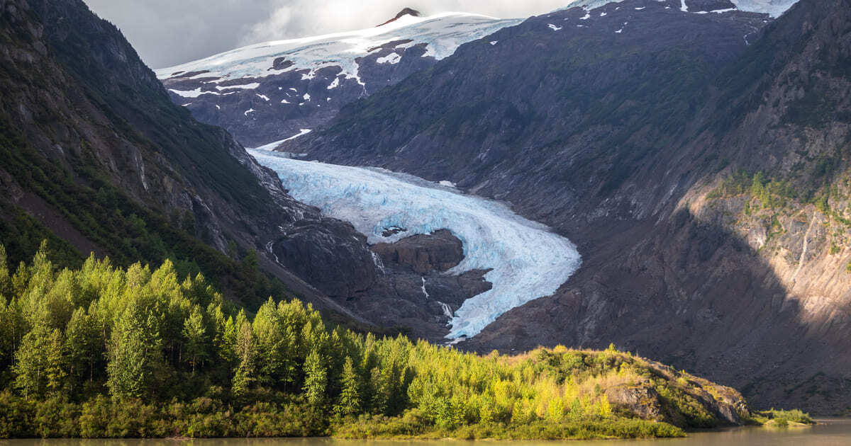 Glacier descending between rocky peaks, fronted by a vibrant green forest.