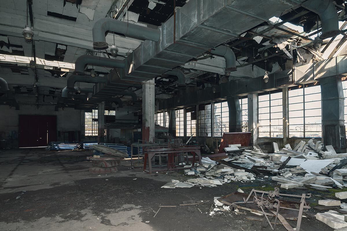 Abandoned industrial hall with debris, pipes, and damaged ceiling.