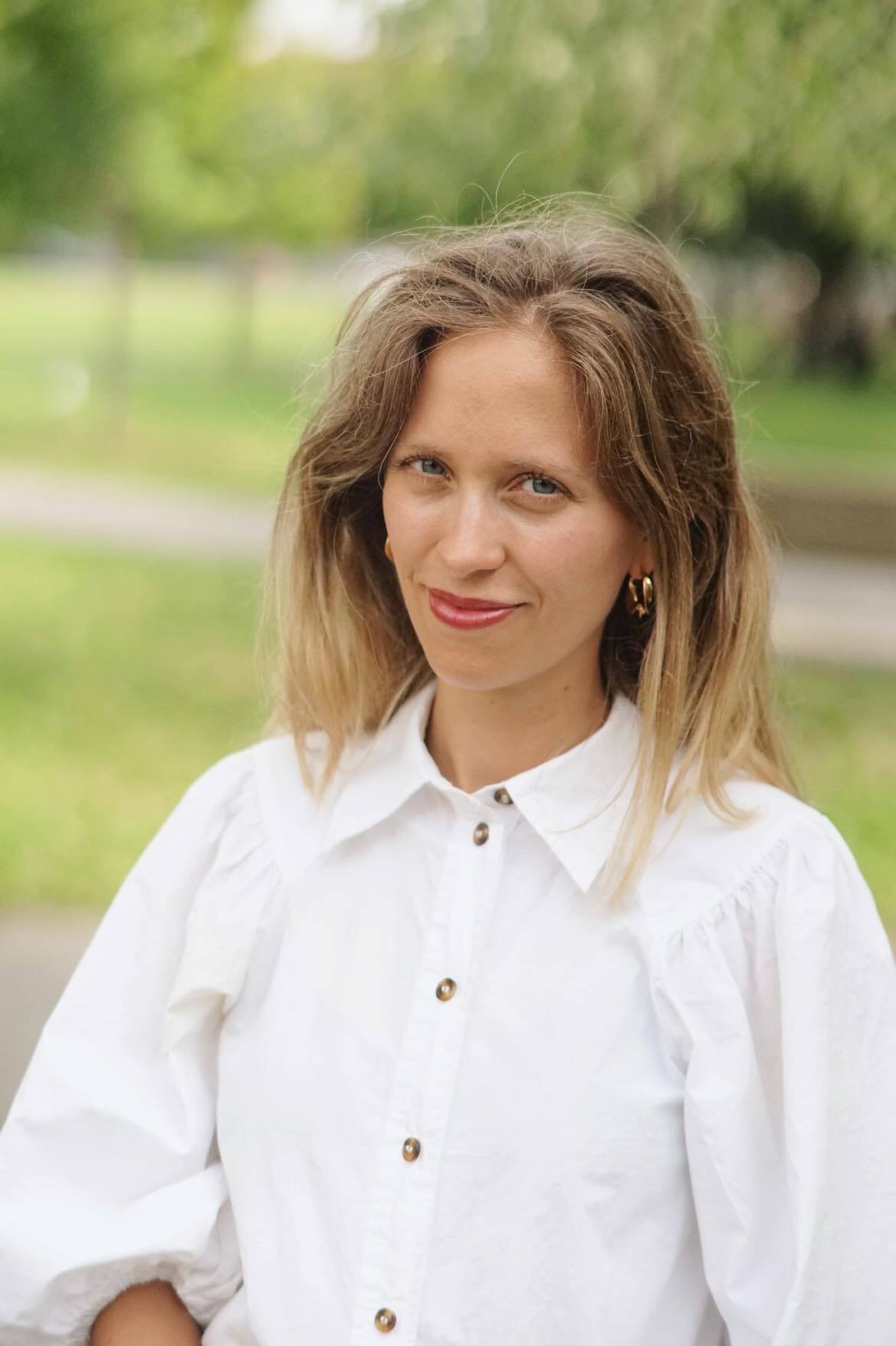 Jeune professionnelle en chemise blanche souriante, ambiance verdoyante et décontractée.