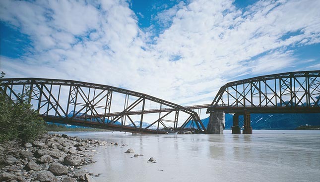Steel truss bridge over a river set against blue skies and mountainous backdrop, showcasing engineering marvels.
