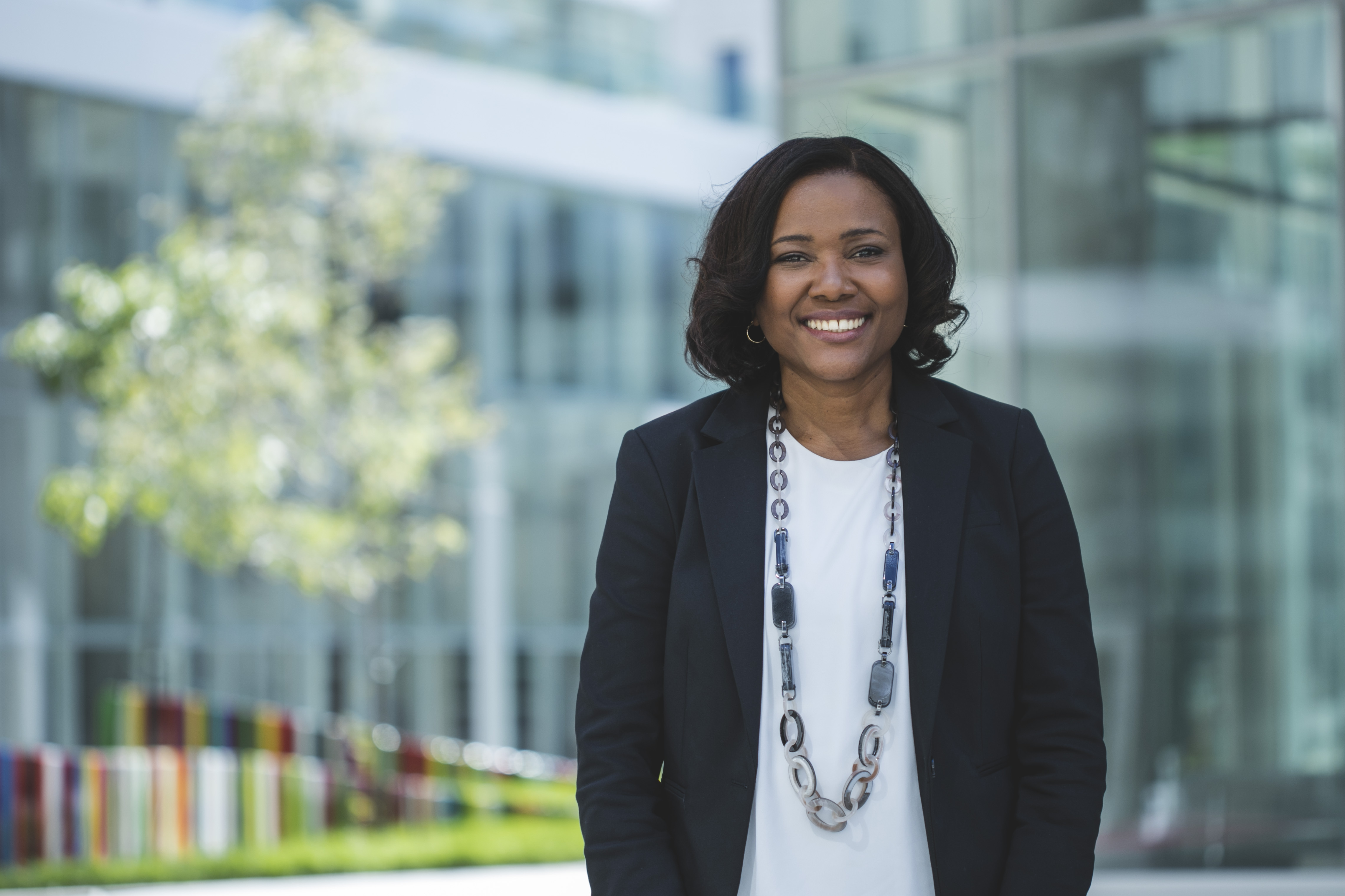 Professional woman smiling outdoors with modern building and greenery in the background.