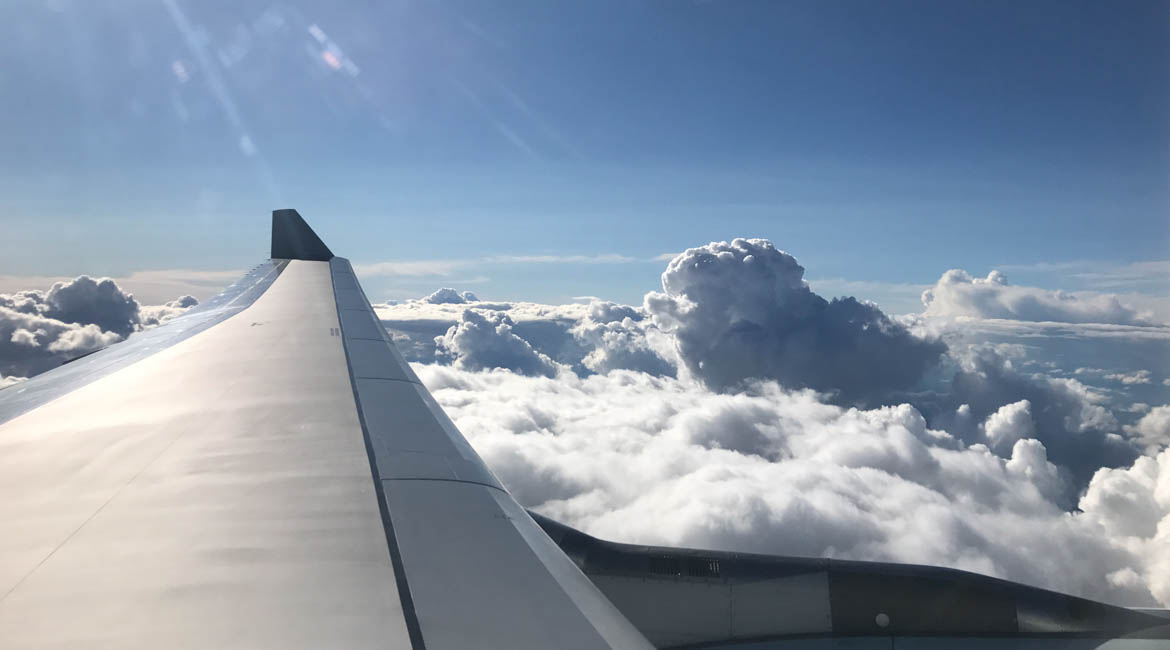 View from a plane window showing wing over clouds with blue sky.